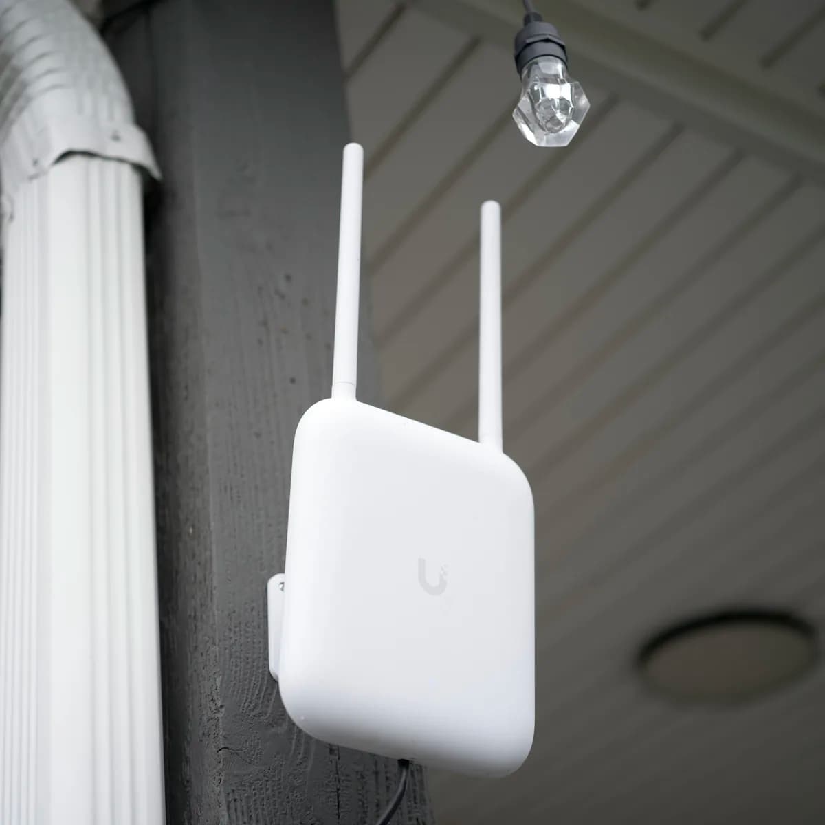 A U7 Pro Outdoor on a porch post under a white soffit, both external antennas fitted, a glass string-light bulb hanging above it.