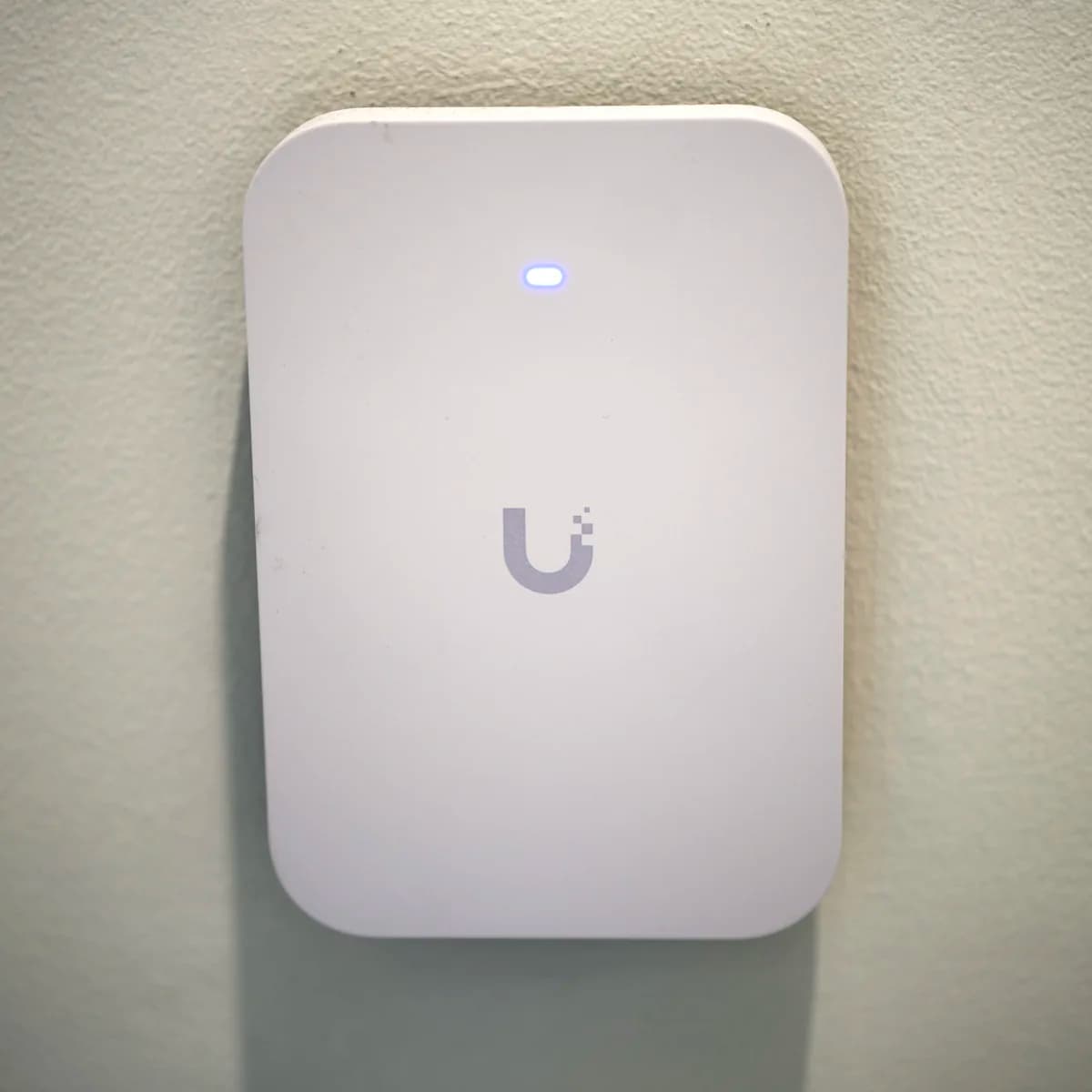 A U7 Pro XG Wall mounted flat on a painted wall, status LED lit blue above the Ubiquiti mark.
